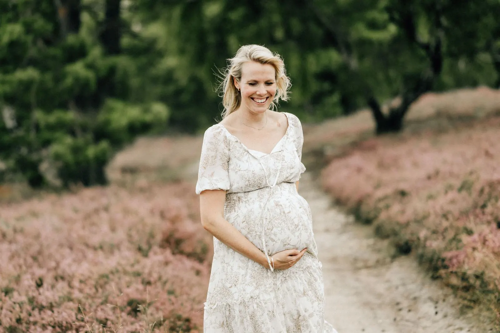 Lächelnde Schwangere im geblümten Kleid auf einem Sandweg in der blühenden Heide