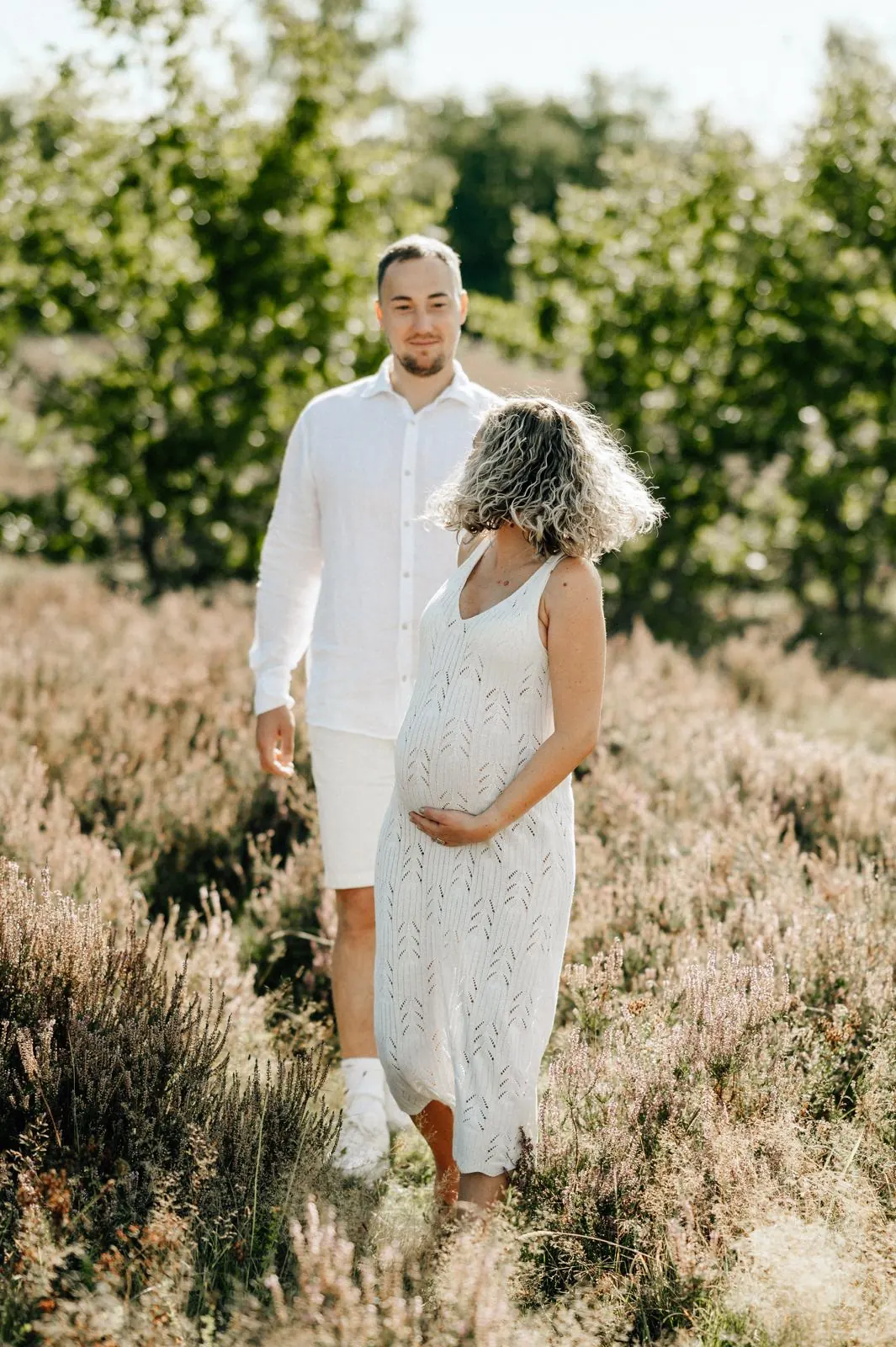 Schwangere im weißen Strickkleid geht durch die Heide, ihr Partner folgt ihr