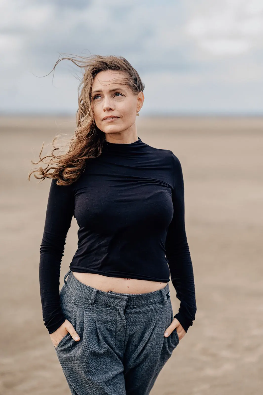 Frau im schwarzen Oberteil am weiten Strand, der Wind spielt mit ihren Haaren