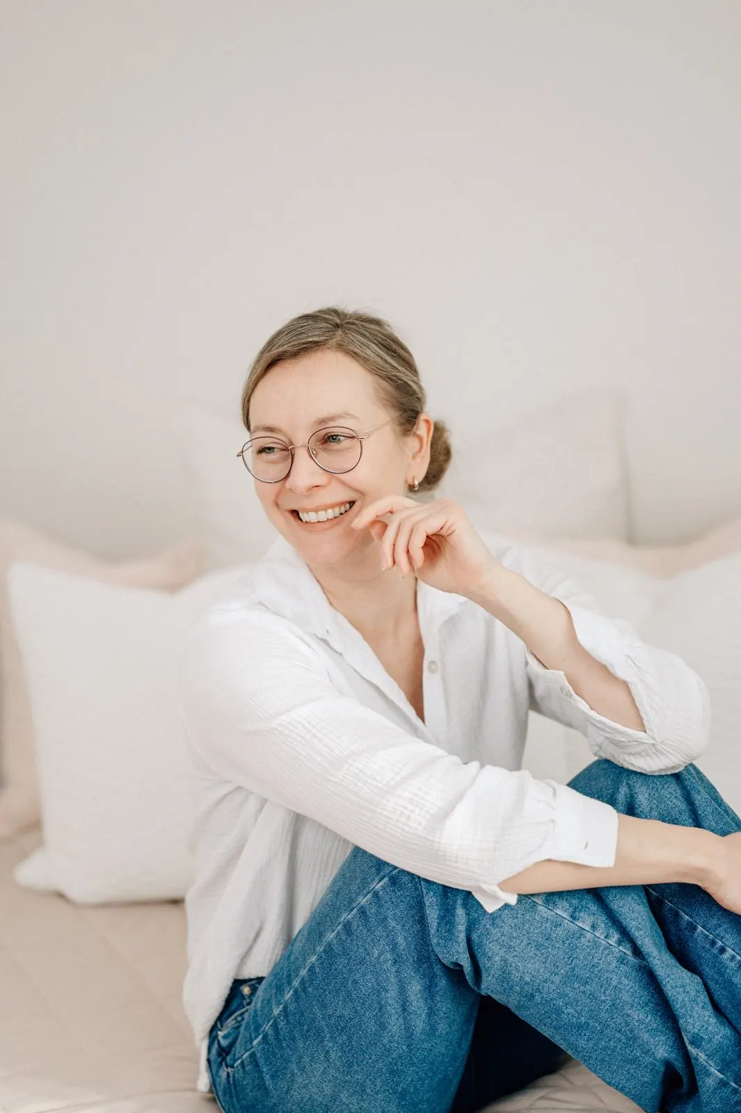 Julia Aßmann mit Brille lacht, im weißen Hemd auf dem Studiobett