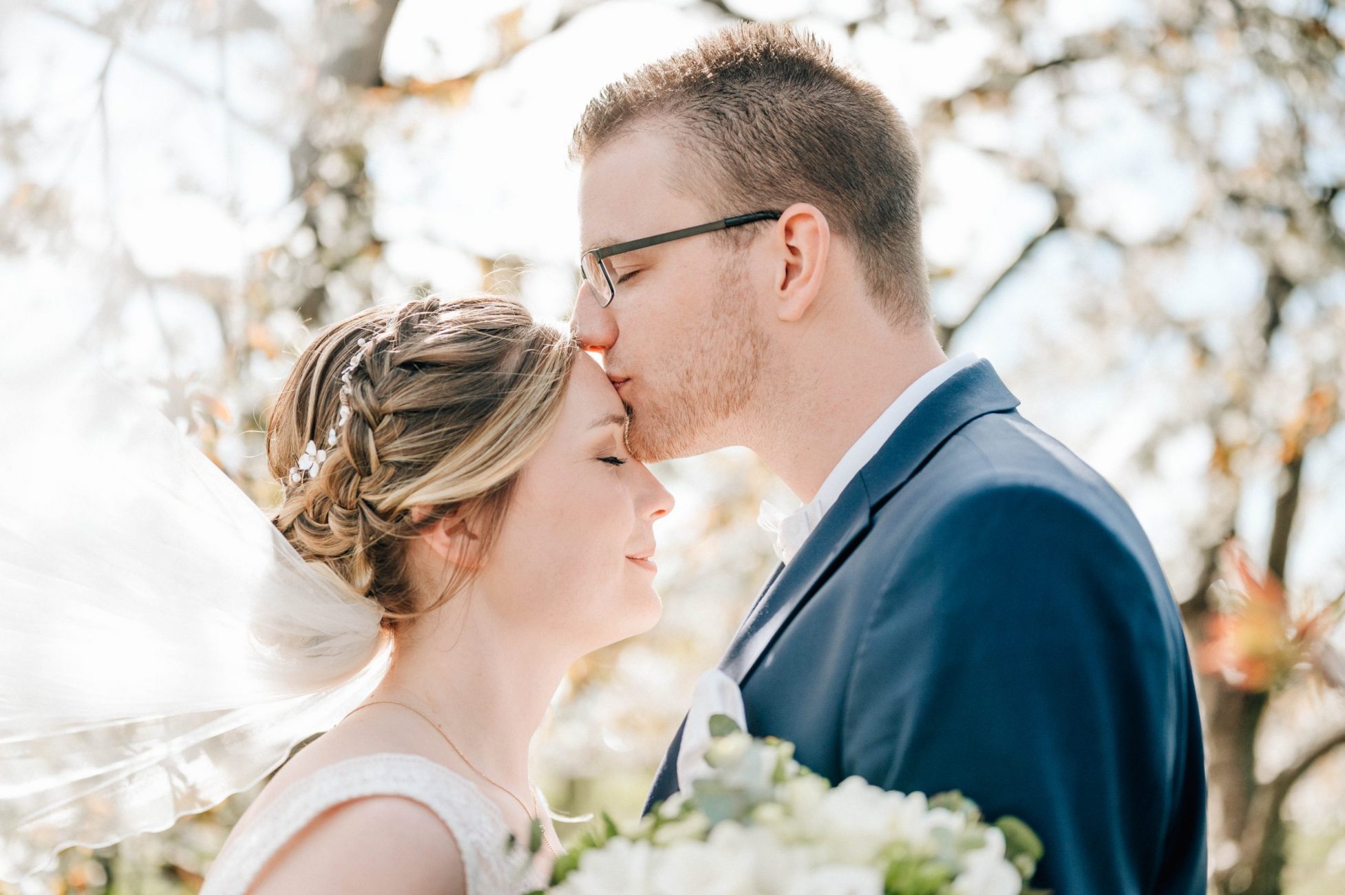Bräutigam küsst Braut auf die Stirn und Schleier der Braut weht im Wind in den Obstblüten beim Paarshooting in Jork am Hochzeitstag